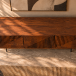 Elevated view of the Rhythm bar cabinet in a warm brown wood finish, with a broad grained top, three doors patterned with horizontal and vertical grooves, and slim dark legs.