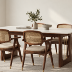 Angled view of the OREN dining table with an ivory top, brown wood finish supports and vertical gold accents, paired with six chairs featuring woven cane backs and beige upholstered seats.