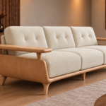 Angled side view of the Grand Turismo sofa in ivory Aston 902 fabric, showing stitched back cushions, a curved light wood side panel, broad raised armrests, rounded base and tapered legs.