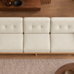 Overhead view of the Grand Turismo three-seater sofa in ivory Aston 902 fabric, showing three equal seat cushions, stitched back cushions, broad wooden armrests and a rounded light wood frame.