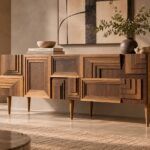 Angled view of the Labyrinth bar cabinet in a warm brown wood finish, showing stepped geometric front panels, a smooth right side with visible grain and slim tapered legs.