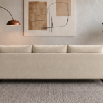 Rear view of the Madeira sofa in cream Amy 904 upholstery, showing a broad patterned back panel, three separate back cushions and rounded side frames in a warm teak finish.