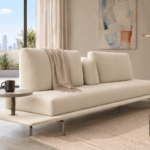 Angled view of the Solaris sofa in ivory upholstery, with a long bench seat, separate back cushions, a central bolster, an extended platform, slim metal legs and a round side table.