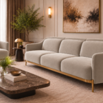 Angled side view of the Duet three-seater sofa and matching armchair in light beige patterned Aston 924 upholstery, showing rounded arms, separate cushions, gold finish base trim and slender legs.