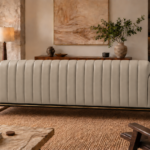 Rear view of the Venetia sofa in light taupe leatherette, showing full-height vertical channels, rounded arm ends, a dark base rail with gold trim and gold-finish metal supports.